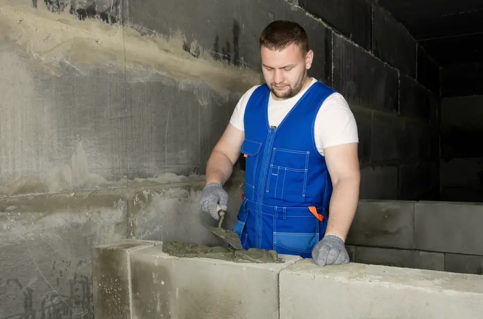 Maçon en salopette qui monte un mur de blocs et garnit les joints au mortier