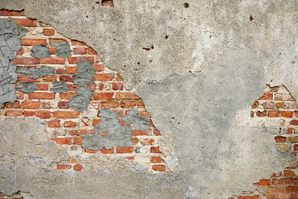 Mur ancien dont l’enduit gris est tombé par plaques et laisse voir la brique rouge